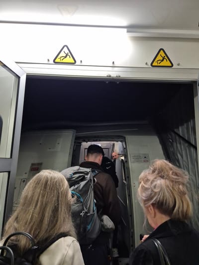 Passengers are disembarking from an airplane. The interior view shows people exiting through the aircraft door. A yellow warning sign with a figure falling is visible on the interior wall. "Salida Exit" is printed next to the emergency exit door. The location is likely at the José María Córdova International Airport in Rionegro, Colombia.