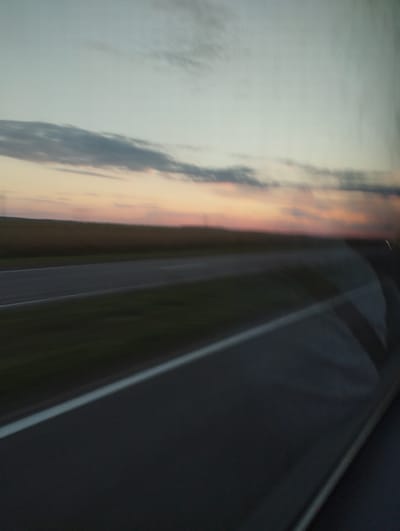 The image depicts a blurred view from a moving vehicle, likely a car, traveling along a road at dusk. The sky shows a gradient of soft pastel colors, with streaks of orange, pink, and pale blue, indicating either sunrise or sunset. Wispy clouds are visible across the sky.
Below the sky, a strip of dark land, possibly fields, stretches horizontally across the frame. In the foreground, a road is visible, characterized by its asphalt surface and a white line marking the lane. The road appears to curve to the right. The overall impression is one of motion, with the blur suggesting a speed of travel.
Given the context provided, the scene is located in the Дукорскі сельскі Савет in Belarus. The image captures a common rural or semi-rural landscape along a road in this region, characterized by open fields and a sky that transitions through the diurnal cycle. There are no discernible people or specific architectural structures in the image due to the blur and the focus on the natural landscape and the road. The environment is open and appears to be unpopulated at the immediate roadside, typical of a transit corridor through agricultural or undeveloped land.