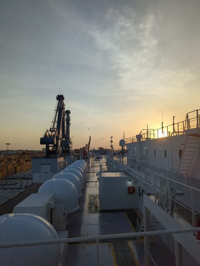 The image shows a sunrise at the port of Hambantota, Sri Lanka. Two large cranes are visible in the distance, along with rows of stacked shipping containers. In the foreground, a large white ship is docked. The ship has a white railing along its deck and several large, white, rounded structures that appear to be life rafts or storage units. The sky is a pale orange and yellow, with scattered clouds. The sun is low on the horizon, casting a warm glow over the scene. The overall impression is one of a busy industrial port at the beginning of a new day.