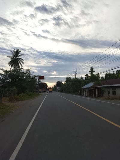 The image depicts a rural road scene in Meunasah Kumbang, Indonesia, likely during the early morning or late afternoon given the diffused light and cloudy sky. A paved road with a white line marking the left lane and a yellow line marking the right lane stretches into the distance. The road is flanked by lush green vegetation and trees on both sides. On the left, a tall palm tree stands prominently. A few simple buildings with corrugated roofs are visible on the right side of the road, appearing somewhat aged. A utility pole with overhead wires is present on the right. In the distance, a yellow truck and a motorcycle are traveling on the road, indicating ongoing activity. A red traffic sign with white lettering is mounted on a pole on the left side of the road. The sky is filled with dramatic, layered clouds, with hints of lighter sky visible in the distance, possibly indicating the setting or rising sun. The overall atmosphere is calm and rural.