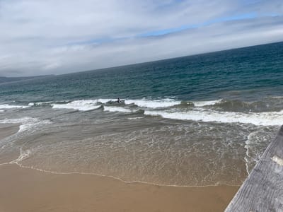 The image depicts a wide view of the ocean and a sandy beach in Hermosa Beach, California, United States of America. The sky is partly cloudy with streaks of blue sky visible through the overcast. Gentle waves are breaking on the shore, leaving a foamy white line on the wet sand. In the middle distance, several people are in the water, some appearing to be surfing or bodyboarding. The ocean water is a deep teal color, and the horizon line is clearly visible. To the right of the frame, a portion of a wooden pier or boardwalk is visible, adding a textural element to the foreground. The overall scene is one of a typical, pleasant day at the beach.