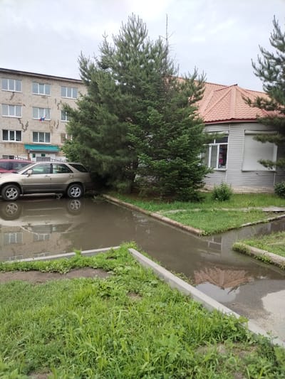 The image depicts an outdoor scene in Pokrovka, Russia, likely during the daytime given the ambient light. The weather appears overcast, with a dull sky suggesting a cloudy day. The setting is a residential or public area, characterized by a brick building with multiple windows, a smaller building with a red tiled roof and siding, and mature evergreen trees. In the foreground, a significant puddle of water reflects the surrounding environment, indicating recent rain or poor drainage. A beige SUV is parked near the trees, partially submerged in the water. A portion of another vehicle, possibly red, is visible on the far left. The grounds are covered in green grass, with some areas showing signs of cultivation or planting beds. Concrete curbing defines the edge of the paved area and the grassy sections. One of the buildings displays a Russian flag hanging from a window. The overall impression is one of a mundane, possibly damp, day in a Russian locality. There is no discernible text within the image.