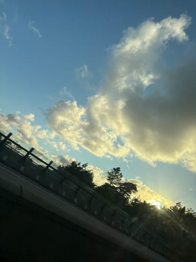 This image depicts a sky with scattered clouds, illuminated by the setting sun, seen from the perspective of a vehicle traveling on a road. The road, which is angled diagonally from the bottom left to the top right of the frame, is bordered by a dark barrier. Beyond the barrier, a dense line of trees is visible, with the sun's rays piercing through the foliage. The sky is a mixture of blue and white, with dramatic clouds of varying sizes and densities. The lighting suggests it is late afternoon or early evening. The location is identified as Ödsmål, Sweden, implying a scenic natural environment adjacent to a roadway. There are no people or specific identifiable structures visible in the image besides the road infrastructure and trees.