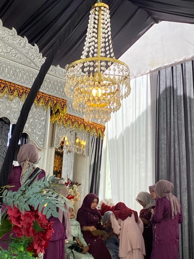 This image captures an indoor event in Matangbaroh, Indonesia, featuring an ornate decorative setting and several women. The upper portion of the frame displays a black fabric ceiling drape on the left, meeting a white fabric tent panel on the right. A large gold-toned chandelier, with multiple tiers of illuminated clear crystal or glass ornaments, hangs centrally. Below the black draping, elaborate white carved panels, featuring intricate floral and geometric designs, form a decorative backdrop on the left, topped by a gold and red patterned valance with hanging tassels. A smaller, multi-bulb chandelier is visible further back within the decorated area. On the right, vertical white and dark grey pleated curtains are visible.

In the foreground, a substantial floral arrangement, comprising prominent red flowers and green eucalyptus-like foliage, partially obscures a woman in a purple outfit and light purple hijab, seen from behind. To her right, a woman in a light green tunic and matching hijab is seated, looking towards the right, with a black bag visible on her lap. On the right side of the frame, a group of women, predominantly in purple or maroon outfits and various colored hijabs, are gathered. One woman, dressed in a light pink top and maroon hijab, is bending forward with her back to the camera. Adjacent to her, a woman in a maroon outfit and grey hijab stands, looking downwards. Further right, a woman in a purple outfit and light purple/grey hijab stands, appearing to hold a small object in her hands. A fourth woman, in a similar purple outfit and light grey hijab, is partially visible on the far right. All women present are wearing headscarves.
