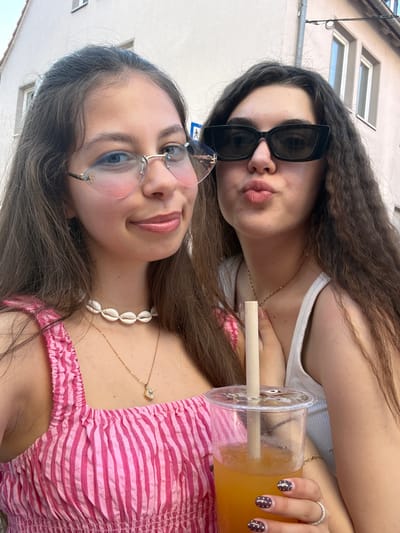 Two young women pose for a selfie. The woman in the foreground wears pink-tinted glasses, a pink and white striped top, and a shell necklace. She is smiling. The woman in the background wears black sunglasses, a white tank top, and a necklace. She is making a kissing face. Between them is a clear plastic cup filled with an orange liquid, and a wooden straw. The woman in the background is holding the cup, and her hand has a ring on it, and her fingernails are dark brown with white dots. The background shows a light-colored building with white-framed windows. This image was taken in Hanau, Germany.