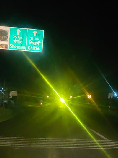The image depicts a night scene on a road, likely in or near Deulghat, India, as indicated by the text on a green directional sign. The sign shows two directions: "75 Km. Shegaon / शेगाव" and "31 Km. Chikhli / चिखली", both with upward-pointing arrows. The road has visible lane markings and reflective markers. The scene is illuminated by bright headlights, causing significant glare and lens flare that obscures details of the environment and any vehicles or people present. There are also smaller orange directional signs with curved arrows visible further down the road. The overall impression is one of a dimly lit highway at night, with artificial light sources being dominant.