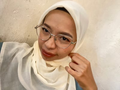 A woman wearing a cream-colored hijab and silver-rimmed glasses poses for a selfie against a textured, off-white wall. She has on a cream-colored hijab draped around her head and neck, and a light-colored scarf that is also visible. Her makeup includes dark eyeliner and reddish-brown lipstick. She is wearing a blue garment underneath her hijab and scarf. Her right hand is brought up to her neck, adjusting the hijab or scarf. The background appears to be an indoor environment, possibly a cafe or a home, with the wall showing some imperfections and slight discoloration. The lighting is soft and diffuse, casting a gentle illumination on her face. The overall impression is that of a casual, personal portrait. There is no indication of the specific location within Kembang Harum, Indonesia.