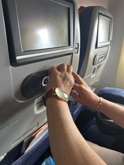 The image shows a close-up view of an airplane cabin. In the foreground, a person's arm wearing a gold-faced watch with a brown leather strap is visible. Their hand is touching a control panel on the back of an airplane seat. The control panel has a screen and several buttons. Text in Cyrillic and English on the panel reads "Пульт должен быть убран" and "Stow and Latch Handle". Another hand, wearing a delicate gold bracelet, is also visible reaching towards the control panel. The background shows the backs of airplane seats, with one seat featuring a small screen. The environment appears to be a typical airplane interior, with blue upholstery and grey plastic elements. There is no indication of the city of Nochvino, Russia, within the image.