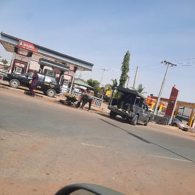 The image captures a roadside scene in what appears to be a sunny, daytime setting, likely in Nigeria given the context. The photograph is rotated approximately 90 degrees clockwise.
In the foreground and mid-ground, a gas station with a distinct awning is visible. The station features multiple fuel pumps. Visible text on the station's signage includes "SHUMA OIL & GAS LTD." and the "TOTAL" logo. A dark sedan or small car is parked at one of the pumps, and a person dressed in dark clothing is standing beside it. On the side of this car, text partially reads "SWANK JOYCE."
Further up the road, another vehicle, which resembles a utility or police patrol van with its rear open, is also visible. Near the gas station, a person is bent over, attending to items on a small, wheeled cart or trolley. The road surface is visible, appearing to be paved and somewhat dusty.
The background shows power poles with lines extending across the clear, bright blue sky. A tall, slender tree stands prominently in the middle distance. Buildings with light-colored walls and reddish roofs are also scattered in the background. The right edge of the image shows a partial view of what appears to be a car's side mirror, suggesting the photo was taken from inside a vehicle. The overall impression is one of a typical commercial roadside area under bright, clear weather conditions.