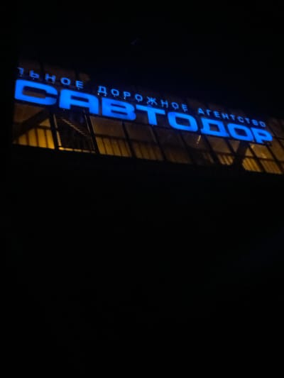 At night, a blue neon sign vertically displays the Russian text "СВЕТОДОР" and above it, "ДОРОЖНОЕ АГЕНТСТВО," indicating the location is likely the office of a road agency in Ижевск, Russia. The sign is affixed to the side of a building with a visible grid structure on its facade. The overall scene is dark, with most of the image dominated by black space, suggesting a nighttime setting.