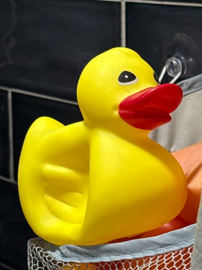 This is a close-up, low-angle shot of a yellow rubber duck toy, possibly a squeaky toy, with a red comb on its head, suggesting it may be designed to resemble a chick. The duck is positioned vertically and appears to be attached to a dark, possibly tiled surface, with a suction cup visible in the background. Part of a mesh bag or container with an orange grid pattern and blue trim is visible to the right of the duck, hinting at a bathroom or storage setting. The lighting is somewhat dim, with highlights on the glossy surface of the duck. The image is sharp, focusing on the toy's features, including its black eye with a white pupil and the textured details of its red comb and body. There is no visible text in the image.