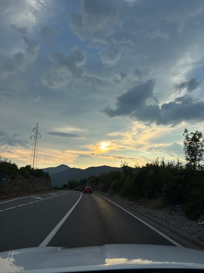 The image captures a view from a vehicle traveling on a road in Liješnje, Montenegro, likely during sunset. The sky is filled with dramatic clouds, with the sun partially obscured by them, casting a warm, golden light. The road ahead curves to the right, marked by white lane lines. On the left side of the road, there's a steep, rocky embankment covered in greenery, and a tall electricity pylon stands prominent. The right side of the road is bordered by dense vegetation and trees. Several cars are visible on the road, moving in the same direction as the viewer. In the distance, mountains form a silhouetted backdrop against the colorful sky. The hood of the vehicle from which the photo is taken is visible in the foreground.