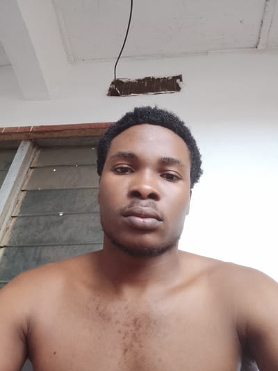 The media file is a close-up, chest-up photograph of a young Black man. He is shirtless, and his skin tone is a warm brown. He has short, dark, curly hair and a light stubble on his chin and upper lip. His expression is neutral, with his gaze directed forward.

The background is softly lit. To the left, there is a window with horizontal slats, suggesting it might be a jalousie window, common in some tropical regions. The window frame appears to be made of wood. Above the window, there is a reddish-brown band, possibly part of the building's exterior trim. The wall behind him is painted white. In the upper portion of the image, a dark wire hangs from the ceiling, and there is a rectangular opening in the ceiling, revealing some dark material.

The lighting suggests it could be daytime, possibly indoors or in a shaded outdoor area, given the lack of harsh shadows. The overall impression is casual and personal. The setting, with the window and building style, along with the provided location context of Orodo, Nigeria, suggests a residential or semi-outdoor environment in that region. There is no visible text in the image.