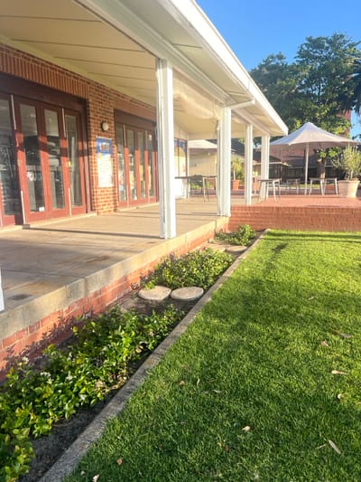 The image displays an outdoor commercial or recreational setting in Applecross, Australia. The scene is dominated by a red brick building with a covered veranda extending along its length. The building facade features multiple wooden-framed glass double doors and an exterior wall-mounted light fixture. Posters are affixed to the brick wall and visible through the glass doors.
The covered veranda has a concrete floor, a white awning supported by white pillars, and translucent roll-up blinds visible along its outer edge. Two tall, narrow tables with dark bases are positioned on this covered veranda.
Separating the veranda from a vibrant green lawn is a narrow garden bed. This bed contains green foliage plants, some exhibiting small white flowers, and two round, light-colored embedded discs. The lawn itself contains scattered dry leaves, and distinct shadows across it indicate low-angle sunlight.
Beyond the lawn, an additional paved outdoor seating area is visible. This area features multiple square-topped tables with accompanying dark chairs, shaded by a large light-colored umbrella. A large potted plant is also present in this section. In the background, other buildings with terracotta roofs and mature green trees are discernible. No individuals, actions, or direct interactions are visible in the scene.