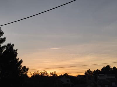 The image presents a landscape view taken in Sant Esteve Sesrovires, Spain during a sunset. The sky transitions from greyish blue at the top to orange near the horizon, with visible contrails from airplanes cutting across the sky. Silhouetted trees and the outlines of buildings form the lower portion of the frame. Power lines stretch across the top and middle portions of the view. A large dark tree appears on the left side.