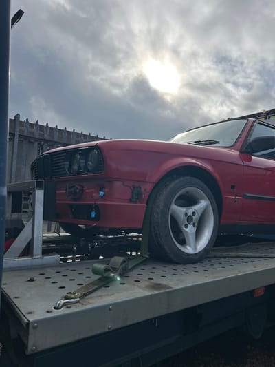 A red car is secured on a flatbed tow truck against a backdrop of a cloudy sky with the sun breaking through. The car is facing downwards and slightly to the left. The front of the car is visible, including the grill, headlights, and front wheel, which has a silver, three-spoke rim. Ratchet straps are used to hold the car in place on the metal bed of the tow truck. The location is presumed to be Rutherglen, United Kingdom. No people are visible in the image.