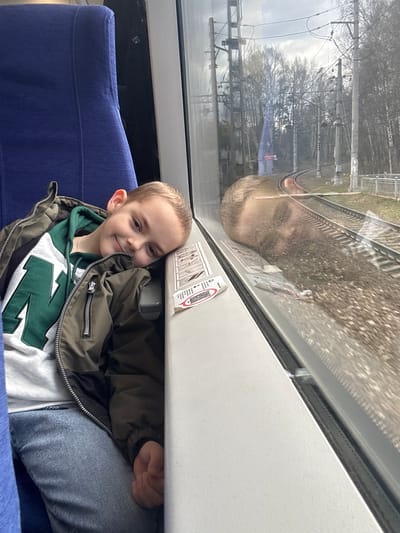 A boy is sitting inside a train car. He is wearing a green and white sweatshirt with a large "N" on it, a brown jacket, and jeans. He is smiling. A blue seat is visible. Outside the train window, a railway track, trees, and overhead power lines are visible. The exact location cannot be determined from the image alone.