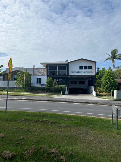 This image presents a daytime street view, likely in Riverhills, Australia, focusing on a modern building situated alongside a road. The building features a multi-story design with a white upper facade and a darker, possibly dark grey or black, lower section. Prominently displayed on the white section is the text "Nurturing Curiosity," while the darker area below shows "Eden Academy" accompanied by a stylized leaf or plant logo. Multiple rectangular windows are visible on both sections of the building.

In front of the building, a paved footpath runs parallel to a two-lane asphalt road with a solid white line marking the edge and a dashed white line in the center. A yellow line is also visible near the edge of the road. To the right of the road is a wide, grassy verge. Lush green trees and shrubs, including a tall palm tree, are scattered around the property and in the background. The sky above is bright with scattered white clouds.

In the foreground, a yellow diamond-shaped road sign, common for school zones, is visible. It depicts the silhouette of two children and clearly reads "CROSSING AHEAD," indicating a pedestrian crossing point or a school zone. No people or explicit activities are visible in the static scene. The lighting suggests it is either morning or afternoon, with clear weather conditions.