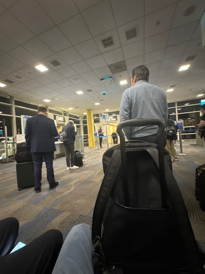 The image depicts an indoor scene at an airport terminal, specifically at Gate D41 and D43A/D43B, identified as Ronald Reagan Washington National Airport (DCA) in Arlington, Virginia, United States. The time of day appears to be night or early morning, indicated by the dark exterior visible through large windows on the left.
The perspective is from a low, seated position, looking forward towards the gates. In the immediate foreground, two pairs of human legs are visible: one set in dark trousers with an illuminated phone screen on the lap, and another set in light blue trousers. Directly in front of the right leg is a black backpack, its carry handle extended, partially obscuring the back of a man wearing a light blue, long-sleeved polo shirt with dark hair.
Further into the scene, towards Gate D41, a man in a dark navy suit and black shoes stands with his back to the camera, positioned next to a black rolling suitcase topped with a smaller black bag. To his right, another man, facing slightly away from the camera, wears dark casual attire, light-colored pants, and white sneakers, with a dark backpack and an adjacent black rolling suitcase. Both individuals appear to be waiting or processing at the gate. Behind them, queuing stanchions with blue retractable belts are visible.
Near Gate D43A/D43B, a man in a purple short-sleeved collared shirt stands, facing the gate counter. Several other indistinct figures are visible throughout the background.
The environment features a carpeted floor with a geometric pattern of blue and beige/brown wavy stripes. The ceiling consists of white grid tiles with numerous square fluorescent light fixtures, ventilation grates, and a security camera with a blue indicator light. A prominent yellow vertical column is visible between the gate areas.