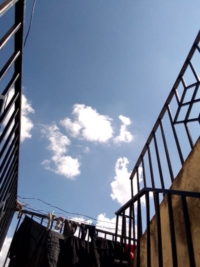 The image, taken in Kalimoni ward, Kenya, provides a low-angle view of a bright blue sky with scattered white cumulus clouds. The foreground is framed by structural elements. On the left, a dark-colored metal railing with vertical bars ascends diagonally. On the right, a similar dark-colored metal railing, incorporating both vertical bars and a geometric grid pattern in its upper section, is attached to a light-colored concrete or stucco wall, indicative of a staircase or balcony. Below the sky and between the railings, a clothesline is visible, from which multiple pieces of dark-colored laundry are suspended, secured by colorful clothespins. A single red fabric item is also present on the clothesline. A large, dark fabric item is draped over a lower, unseen railing or barrier. No individuals are depicted in the scene. The lighting suggests full daylight conditions.