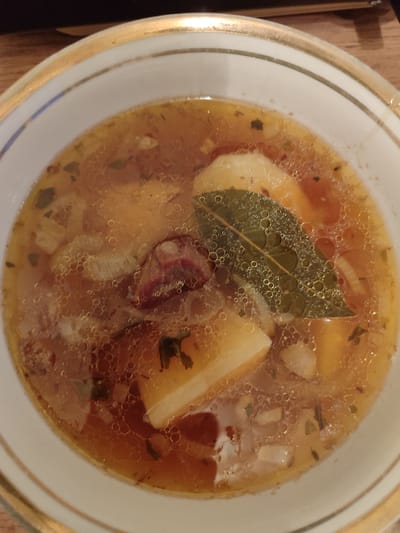 A close-up, overhead shot shows a bowl of soup. The bowl is white with a gold rim. The soup is a light brown color and appears to have chunks of potato, meat, and a bay leaf floating in it. Small bubbles are visible on the surface of the soup. The background is dark and out of focus, suggesting the soup is on a table. The location of Batken, Kyrgyzstan, is not determinable from the image itself.