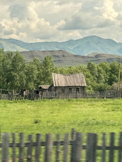 A weathered wooden house with a corrugated metal roof and visible windows stands in a grassy field, bordered by a dilapidated wooden fence. Behind the house, a dense line of green trees forms a backdrop, with layered, muted green mountains and a cloudy sky in the distance. The scene suggests a rural or remote location, likely in the region of Верх-Аскиз, Russia, given the context. The overall impression is one of rustic simplicity and natural beauty.