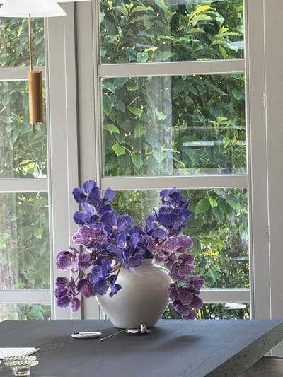 The image depicts a close-up view of a white vase filled with purple orchids. The vase is positioned on the right side of the frame, partially obscuring a dark, textured wall. A thin metal rod extends from the wall, passing through the scene and holding a golden, ribbed object.

Behind the vase and orchids, a large window is visible, divided into multiple panes by white frames. Through the window, lush green foliage is seen, suggesting an outdoor setting. Sunlight filters through the leaves, creating dappled light and shadows. The presence of dense greenery and the quality of light suggest it could be daytime, possibly during pleasant weather.

The overall mood is serene and decorative, with a focus on the vibrant orchids against a backdrop of natural elements and modern interior design. There is no visible text in the image. The setting appears to be an indoor space with a view of a garden or wooded area. The architectural style of the window frames and the interior wall suggest a contemporary home or building.