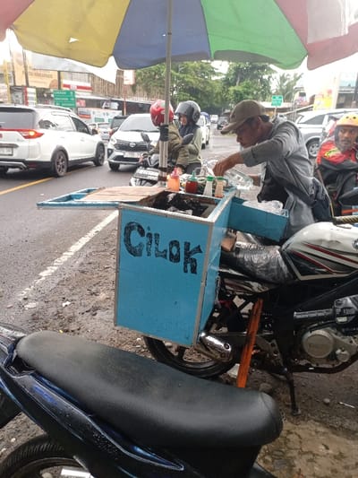 The image shows a street food vendor selling "cilok" (Indonesian meatball snacks) in Kecamatan Caringin, Indonesia, located at latitude -7.523 and longitude 107.485.  The vendor is seated on a black and silver Yamaha motorcycle, which is parked on the side of a wet street. An umbrella, with yellow, green, and blue sections, is set up above a small, blue, wooden cart with the word "Cilok" written in black on it. There are plastic bottles and containers of various sauces on the cart. The vendor is a man wearing a grey shirt and a brown hat. He is using his right hand to hold a bottle of liquid. He is interacting with another person, a woman in a black jacket and grey helmet, who is sitting on the back of a black motorcycle and holding a white plastic bag. In the background, there are multiple cars and other motorcycles parked on the street.