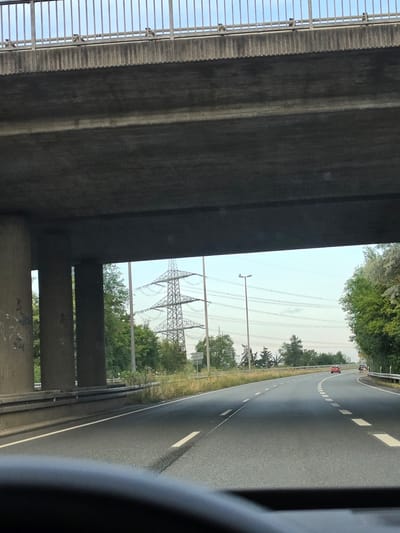 The image captures a perspective from inside a vehicle driving on a highway under an elevated road. The highway has two lanes of asphalt with white dashed lane markings. In the foreground, the dashboard of the vehicle is visible, slightly blurred. The elevated road above is constructed of concrete with metal railings along its edge.

To the left, large concrete pillars support the elevated road, and some graffiti is visible on one of the pillars. Beyond the pillars, there are trees and a large metal pylon with power lines. To the right, more trees line the side of the highway, and a street lamp is visible in the distance.

Several vehicles are present on the highway: a red car is in the right lane ahead, and another vehicle, possibly a truck, is further ahead in the left lane. The sky is clear with a pale blue hue.

The scene is consistent with a typical highway environment in Germany, potentially near an overpass. Based on visual cues like the highway infrastructure, vegetation, and common vehicle types, this scene is likely located in a suburban or semi-urban area. Given the provided information about Rüsselsheim am Main, Germany, this image could be from a section of the A60 Autobahn, which runs through or near Rüsselsheim and features similar infrastructure and landscape.