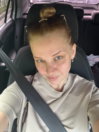 A woman with blonde hair pulled into a bun, wearing sunglasses on top of her head and a light brown t-shirt, is sitting in the driver's seat of a car. She has a purse strap visible over her shoulder and is wearing small hoop earrings. The car's black seat and gray seatbelt are prominent in the frame. The background shows a glimpse of the car's interior and the exterior through the window, indicating it is likely parked or driving in a grassy area, possibly a rural or suburban setting. The image appears to be a selfie taken by the woman.