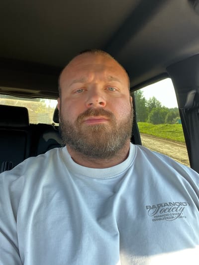 A man is sitting in the driver's seat of a vehicle, looking directly at the camera. He has a shaved head, a full beard with graying hairs, and is wearing a light-colored crewneck sweatshirt with a graphic logo on the left chest. The interior of the vehicle is visible, including the dashboard and a portion of the back seat. The window on the right side of the vehicle shows an outdoor scene with green trees and a dirt road under a bright sky. The location is identified as Старый Кыч, Russia.