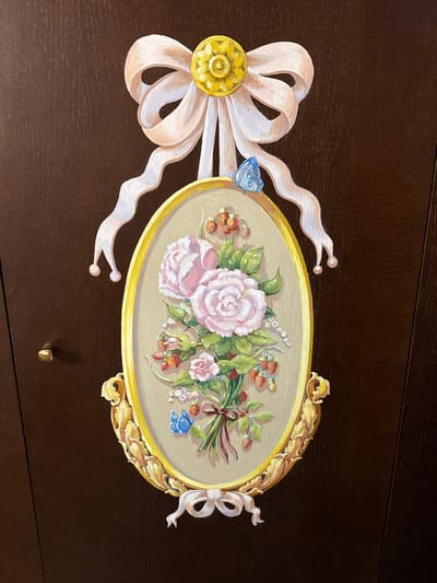 The image displays a painted floral arrangement on a dark brown wooden surface, possibly a door or cabinet. The artwork features a bouquet of pink roses with green leaves and small red berries, all framed within an ornate oval, gilded border. A light pink bow with a golden decorative center is affixed above the oval, with ribbon streamers cascading down. Two blue butterflies are also depicted, one near the top bow and another on the floral bouquet. The dark brown surface exhibits a wood grain texture, and a small, brass-colored knob is visible on the left side of the surface. The image does not contain any recognizable landmarks or context that would indicate its specific location within Ozeretskoe, Russia.