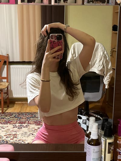 A female individual is depicted in a mirror selfie taken indoors in Popintsi, Bulgaria. The person, with long, dark, wavy hair, is wearing a white short-sleeved crop top exposing the midriff and pink form-fitting athletic shorts. She holds a smartphone with a pink leopard-print case in her right hand, positioned to capture her reflection. Her left hand is raised, with fingers interlaced in her hair. A silver-toned watch is visible on her left wrist, and a beaded bracelet on her right wrist, along with a ring on her left ring finger.
The interior space features a wooden floor partially covered by a multi-colored Persian-style rug with intricate patterns. To the left, a light-colored wooden chair is visible, and behind it, a white ribbed radiator is situated beneath vertical blinds or pleated curtains in beige and white hues. The wall behind the curtains appears light-colored. To the right, behind the individual, a grey and black office chair is present, with a white, long-sleeved jacket draped over its back. A light yellow wall is visible in the reflection to the right. On a dark wooden shelf unit to the far right, a black object (possibly a hat) rests on the top shelf, and a striped object (possibly zebra print) is on a lower shelf. The bottom edge of the mirror reflects a dark wooden surface on which several cosmetic or hair product bottles are placed; one bottle explicitly shows "IKAROV Hair Growth Stimulant," and another partially visible label indicates "L'OREAL ELS." Two pink, circular objects are also on this surface to the lower left.