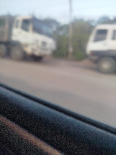 Through a blurry window, two white trucks face each other on a road. The truck on the left appears larger and further away, while the one on the right is closer but partially cut off by the image's edge. Greenery can be seen between and behind the trucks. The foreground is dominated by a dark, textured interior element, possibly part of a car's interior. The overall image quality is soft and indistinct.