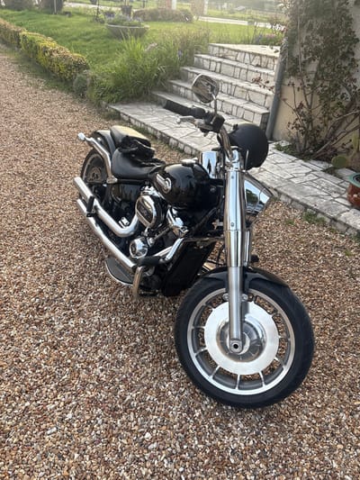 A black cruiser-style motorcycle is stationary on a brown and beige gravel driveway in Bauzy, France. The motorcycle features chrome accents on its dual exhaust pipes, engine components (including a "100" and "V-TWIN" emblem), handlebars, mirrors, and extended front forks. A black helmet rests on the rear passenger seat. The front wheel has a silver multi-spoke alloy rim with a prominent central cap. To the left of the motorcycle, a manicured green lawn is bordered by a trimmed hedge. To the right, a series of light-colored, irregular stone steps ascends alongside a wall with climbing plants and a potted cactus in a terra cotta container. The scene is illuminated by natural sunlight, casting soft light across the foreground. No individuals are present.