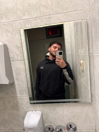 A man is taking a selfie in a bathroom mirror. The bathroom has tiled walls with a marble-like pattern. There is a sink, a soap dispenser, and a chrome faucet visible. The mirror reflects a small glimpse of another room with a red object. The location cannot be determined as Trilistnik, Bulgaria as the details are not discernible.