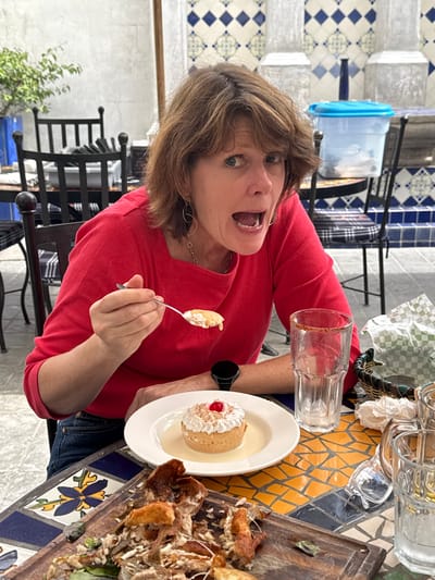 A woman in a red shirt holds a spoonful of custard while standing over a table. A dessert is on a white plate, topped with whipped cream and a cherry. An empty glass, a glass beer mug, and what appears to be the remnants of a meat dish are on the table, which is covered in multi-colored geometric tile. Iron furniture is visible in the background.