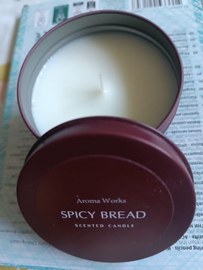 The image shows an open scented candle in a round, burgundy tin. The candle itself is white and appears to be made of wax, with a wick protruding from the center. The lid of the tin is placed open next to it, displaying text that reads "Aroma Works SPICY BREAD SCENTED CANDLE." The background is out of focus but appears to be a light blue and white patterned surface, possibly a book or packaging, with some text visible. There are no people or specific time of day or weather cues. The overall scene is a close-up of the candle, suggesting it is either new or recently used. There is a significant amount of text in various languages on the background surface, but it is too blurry to read in detail.