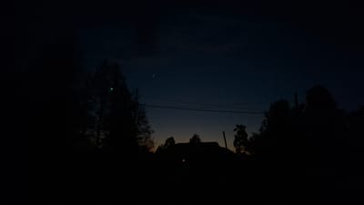 The image shows a dark, likely nighttime, scene in an outdoor environment. Silhouetted trees frame a distant building against a gradient sky that shifts from a dark blue at the top to a light orange/yellow at the horizon. The presence of stars suggests a rural or semi-rural location. Overhead wires are visible. A green light is apparent on a tree. A red light is visible on the building, potentially indicating a light source like a porch light. The foreground is dark and obscures any potential details. While the prompt indicates this image is from Sheshurga, Russia, there is no direct evidence of this in the picture.