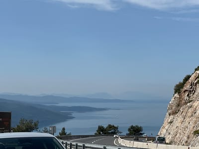 The image presents a scenic view from a highway in Ula, Turkey. The foreground features a white car, partially visible, and a gray metal guardrail along the road's edge. A highway curves, with vehicles visible in both directions. The right side of the image showcases a rugged, light-colored rock face with some vegetation.

In the midground, a body of water extends into the distance, with a series of layered, hazy mountains on the horizon. Power lines traverse the scene, spanning across the water.

The background consists of a clear blue sky with a few wispy clouds. A sign for furniture company 'KOYUNLU' with a partial phone number is located on the left.