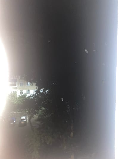 The image appears to be a nighttime or dusk view of a residential area. On the left side of the image, a multi-story building is visible, with windows that suggest interior lighting. In front of the building, there is a parking area with two cars. One car is dark blue, and the other is silver. Trees and foliage are interspersed throughout the scene, obscuring parts of the building and the parking area. The overall lighting is dim, with a bright light source on the far left of the frame, possibly a street light or a strong artificial light. The image is grainy and somewhat blurred, consistent with a low-light photograph. The setting is an urban or suburban residential environment.