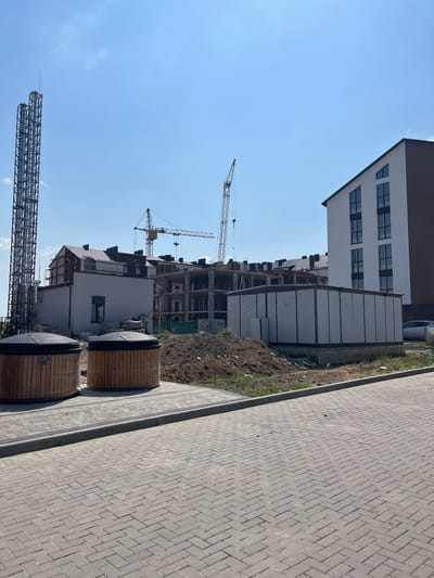 The image captures an outdoor scene in the city of Strumivka, Ukraine, under a clear blue sky with sparse clouds. The foreground features a paved area with interlocking grey pavers arranged in a herringbone pattern, bordered by a concrete curb. To the left, two large, cylindrical wooden structures, wrapped in clear plastic, are positioned on the pavers. Behind these structures, a tall, dark metal industrial tower with a lattice structure stands prominently.
In the mid-ground, a construction site is evident. Several buildings are in various stages of development. One building on the right is a multi-story structure with a white facade and dark brown accents, featuring several angled windows. To its left, a long, low, light-colored industrial building with multiple panels and warning signs is situated. Behind these, more unfinished buildings with exposed concrete and scaffolding are visible. Two yellow construction cranes are present on the site, one in the mid-ground and another further back. A large pile of dark soil or construction debris is located between the wooden structures and the industrial building.
The background consists of more construction and residential buildings, some with visible roof structures and chimneys. The overall impression is that of an active construction zone in a developing area of Strumivka, Ukraine, likely a residential complex. No people are visible in the image.