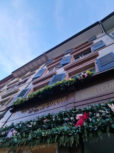 The image shows a building in Rheinfelden, Switzerland. The building has multiple floors with windows that have dark blue shutters. The building's facade features a sign that reads "TRAITEUR CHARCUTERIE". Decorations including garlands and Easter-themed items like pink rabbit figures adorn the building's exterior. The sky above the building is blue with a few light clouds.