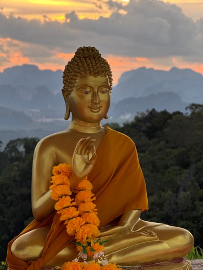 A gold-colored statue of Buddha is seated in a lotus position, adorned with an orange flower garland. The statue is positioned outdoors, with a lush green forest and mist-covered mountains in the background. The sky above displays a dramatic sunset with clouds illuminated by orange and yellow light. The location is identified as Ban Khao Tang, Thailand.