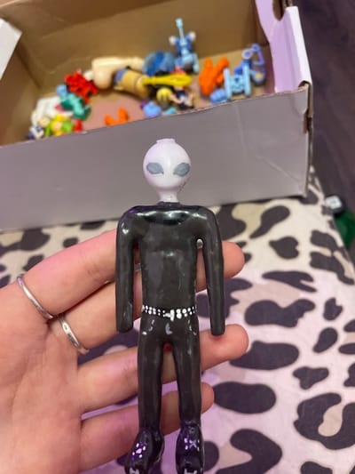 A person's hand with silver rings is holding a black, slender, alien-like figurine with a white head and gray eyes. The figurine is dressed in a black outfit with white dots around the waist and on the boots. In the background, a cardboard box is filled with various small toys, including red, blue, and orange plastic figures, as well as a tan doll. The background surface has a black and white cow-print pattern. The scene is not located in Ladozhskii Trudposelok, Russia.