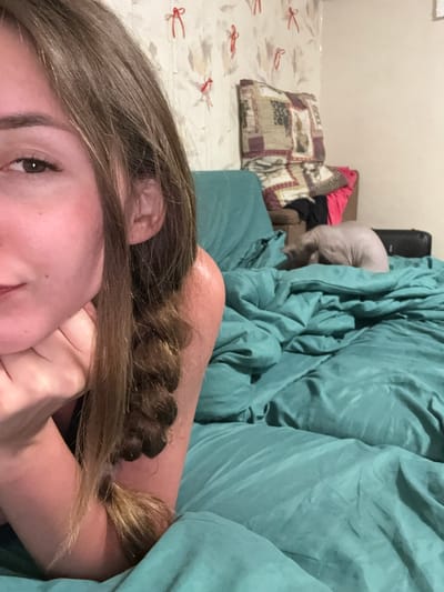 An indoor, close-up, eye-level, slightly angled shot shows a young woman with long brown hair in a braid. Her left arm is bent, with her hand resting on her chin and cheek. She is lying on a teal-colored bed, which is covered with crumpled sheets. In the background, a bald cat is curled up on the bed. The wall behind the bed has light-colored wallpaper with red decorative elements. The room appears to be a bedroom. The image was likely taken in Zhodzina, Belarus.