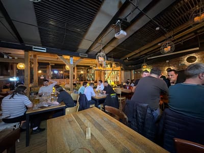 This is an interior, wide-angle photograph of a pub or restaurant located in Sibiu, Romania. The scene depicts a casual social gathering with multiple groups of individuals, predominantly adult males, seated at various wooden tables.
In the foreground-left, a woman wearing a horizontal striped shirt and a man with long hair in a dark t-shirt are seated at a rectangular wooden table with a dark metal frame, each with a large glass beer stein. To their right, a larger, light-colored wooden table occupies the immediate foreground.
In the mid-ground, a group of approximately four men are seated around a square wooden table. One man in a light blue collared shirt faces left, while another in a white t-shirt or hoodie faces the camera with his right hand resting on his head. A third man in a black long-sleeve shirt faces right. Several beer steins and other glasses are on their table. A dark jacket with "MÜNN" visible on the back is draped over a chair in this section.
Further to the right, two men are seen from behind, seated at another table. One wears a dark t-shirt, and the other, further right, wears a dark green t-shirt. Dark jackets are draped over their chairs. Additional patrons are visible in the background, seated at other tables, engaged in conversation.
The establishment features a dark, slatted ceiling with exposed beams and conduits. Multiple spherical, open-frame chandeliers with warm, candle-like lights hang from the ceiling. A white projector is mounted to the ceiling, angled towards the background. Sections of exposed brick wall are visible on the right, decorated with dark, circular elements. Wooden structural posts and beams are prominent throughout the space. Large windows or glass doors in the background reveal blurred exterior lights, indicating it is evening or night. The overall lighting is warm and ambient.