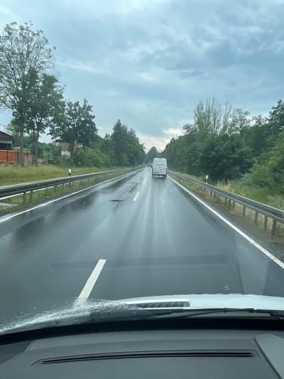 The image shows a forward-facing view from inside a vehicle, looking out at a wet highway. The sky is overcast and cloudy, suggesting rain. The road surface is slick and reflects the sky, with visible tire tracks and puddles. A white van with "Hermes" written on its side is traveling ahead in the right lane. The highway is bordered by a metal guardrail and trees on both sides, with some buildings and structures visible in the background on the left. The lower part of the image shows the vehicle's dashboard and windshield, with water droplets on the glass. The scene appears to be on a highway section outside of Lauterbach, Germany, likely on the Bundesautobahn 26, given the presence of Hermes delivery vans which are common in Germany and the typical highway infrastructure.