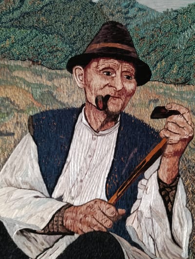 An embroidered artwork depicts an elderly man, likely of Ukrainian descent, wearing a dark hat and a dark vest over a white shirt. He holds a pipe in his mouth and a tinderbox in his right hand. A wooden stick is also visible in his hands. The background shows a landscape with green foliage and hills, possibly representing a rural area. It is presented as an embroidered artwork. The location is identified as Barvinok, Ukraine.