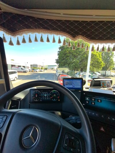 The image shows the interior of a truck cab, looking out through the windshield. The driver's perspective is evident from the large steering wheel with a Mercedes-Benz logo in the center. The dashboard features a digital display showing speed, gear selection (N1), and other vehicle information. A GPS navigation device is mounted on the dashboard, displaying a map and menu options in Russian, including "Search," "My Route," "Map," and "Service."
Outside the cab, a sunny day is suggested by the bright lighting and clear blue sky, with lush green trees visible in the background. Several vehicles, including vans and cars, are parked in what appears to be a commercial or industrial area. A white van with the text "Pneus & Entretien" is parked in front of a building, indicating a tire and maintenance service. The overall scene suggests the truck is parked or stopped in a well-lit outdoor location. The interior of the cab is decorated with a quilted fabric trim along the top of the windshield, adorned with numerous golden tassels, adding a personal touch to the vehicle. The presence of religious icons on the dashboard suggests the driver may be religious. The visible text on the dashboard and GPS indicates a Russian language interface for some of the vehicle's systems, despite the "Pneus & Entretien" sign suggesting a French location context.